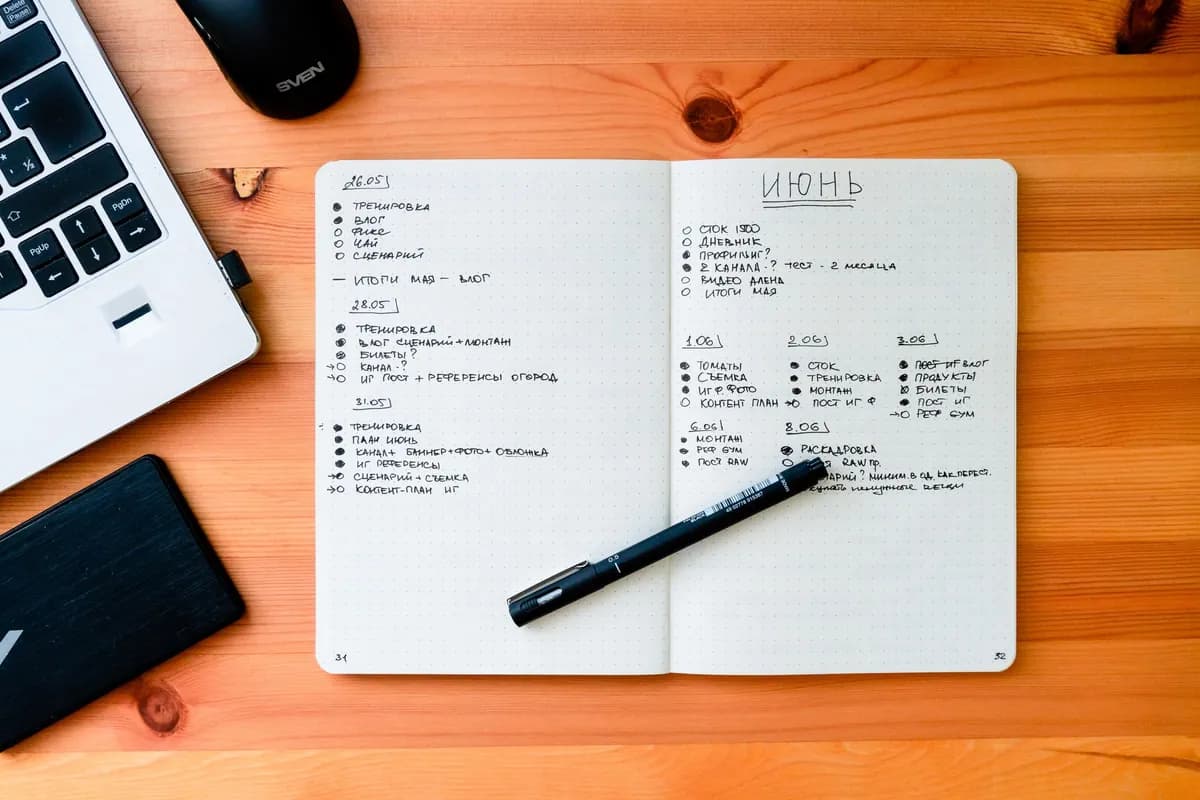 Aufgeschlagenes Notizbuch mit handschriftlichen Listen, Stift und Laptop auf einem Holztisch