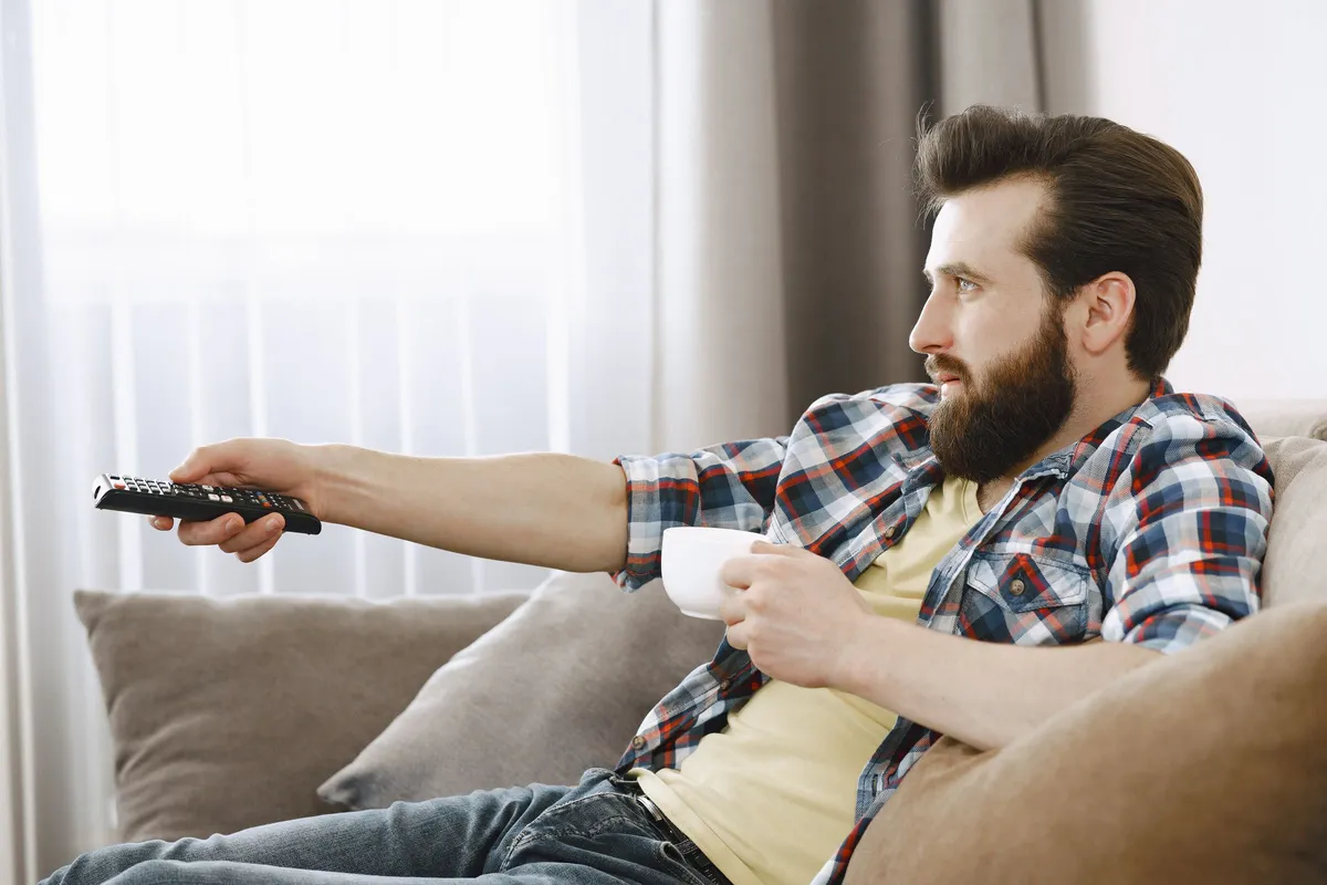 Mann sitzt mit Kaffeetasse auf dem Sofa und richtet die Fernbedienung nach vorn