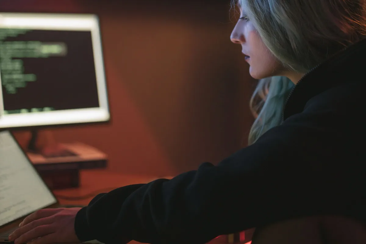 Frau arbeitet abends am Laptop, im Hintergrund ein Monitor mit Programmcode