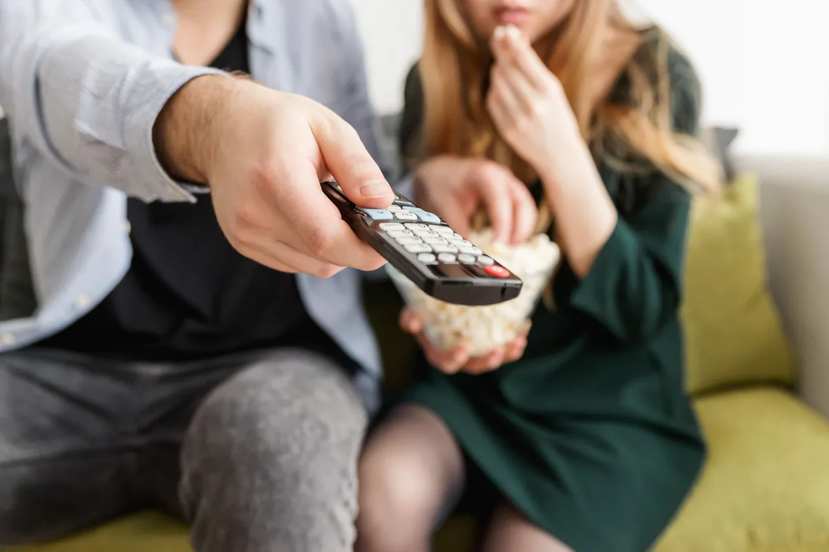 Paar auf dem Sofa mit Popcorn, eine Hand richtet die Fernbedienung auf den Fernseher
