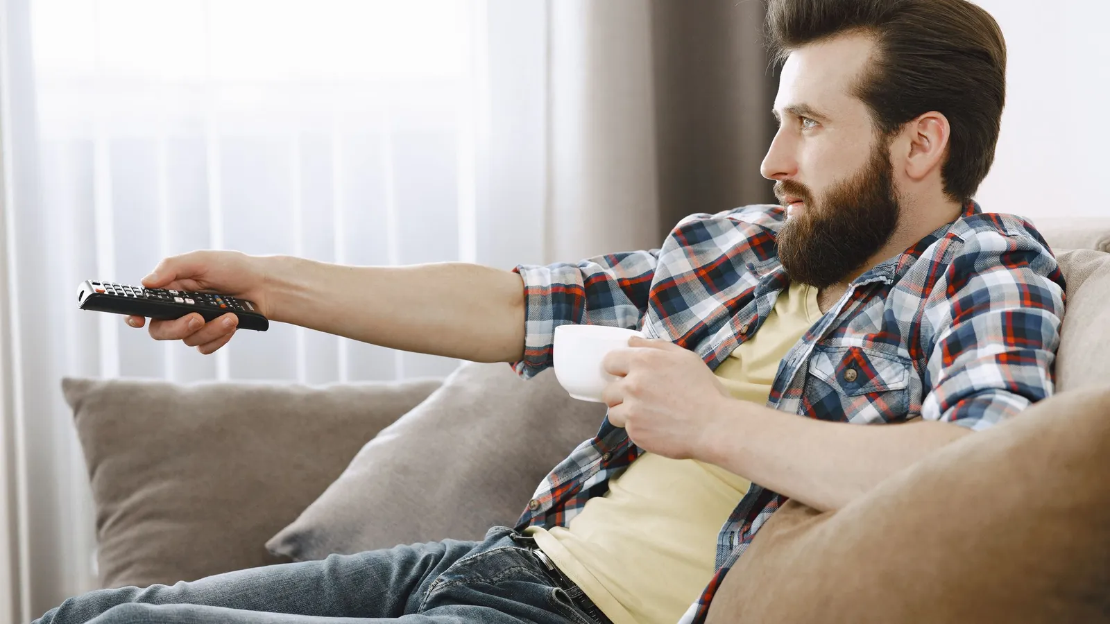 Mann entspannt mit Kaffeetasse auf dem Sofa und richtet die Fernbedienung auf den Fernseher
