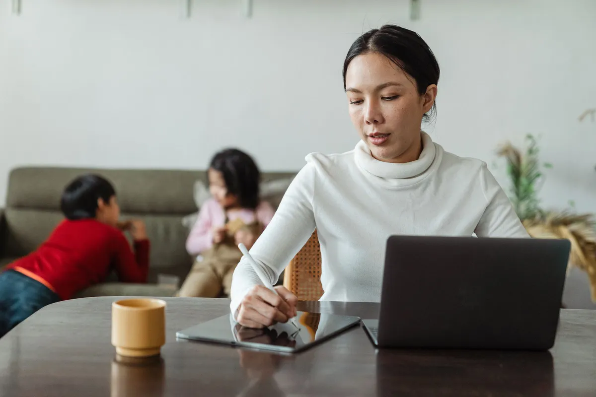 Frau arbeitet mit Tablet und Laptop, im Hintergrund spielen zwei Kinder auf dem Sofa