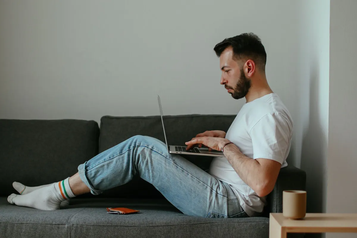 Mann sitzt mit einem Laptop auf einem grauen Sofa