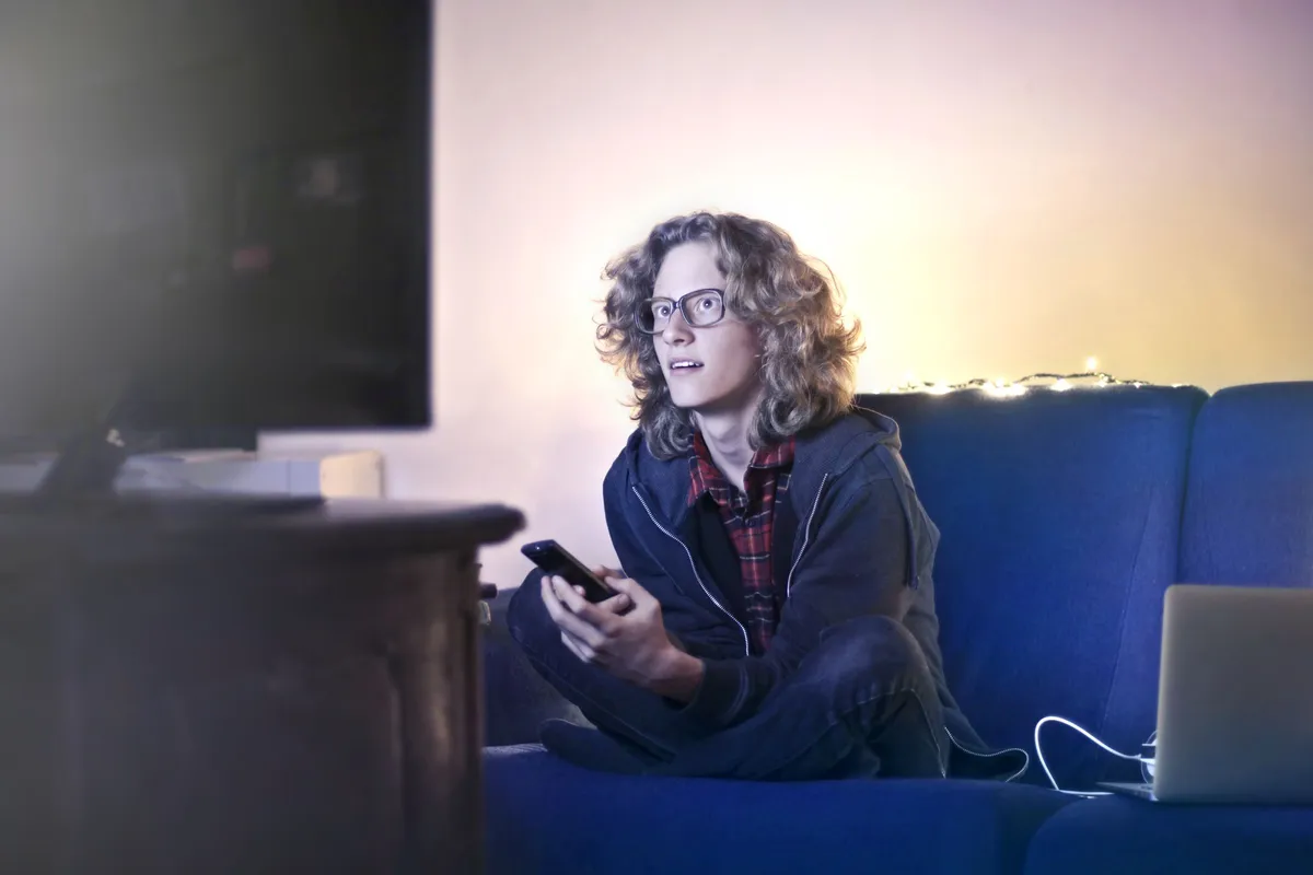 Junger Mann sitzt mit Smartphone auf einem blauen Sofa vor dem Fernseher