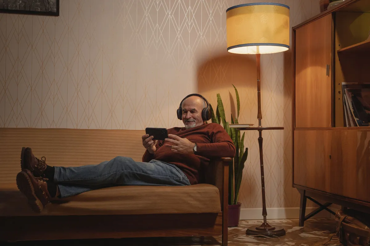 Älterer Mann mit Kopfhörern liegt auf dem Sofa und schaut auf sein Smartphone